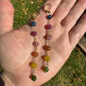 Può includere: Un paio di orecchini pendenti lunghi con ganci dorati a monachella e una catena verticale di perline di pietre preziose sfaccettate multicolori. Le perline sfumano dal blu e rosa al viola, ambra, giallo e verde in una disposizione lineare.