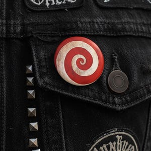 Könnte beinhalten: Nahaufnahme einer schwarzen Jeansjacke mit einem rot-weißen Spiral-Knopf. Die Jacke hat silberne Nieten und einen dunklen Knopf. Aufnäher mit weißem Text sind sichtbar.