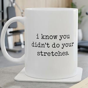 May include: A white ceramic mug with a handle on the left side, featuring the black printed text "i know you didn't do your stretches." The mug sits on a square white coaster, displaying a clean, minimalist design with clear, bold lettering.