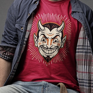 Peut inclure: Un t-shirt rouge orné d'une illustration graphique représentant un visage de diable souriant avec des cornes, un bouc et des lignes blanches rayonnantes. Il est porté sous une chemise bleue déboutonnée à doublure écossaise et un jean bleu.