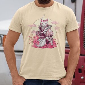 K&ouml;nnte beinhalten: Ein beiges T-Shirt mit einem Aufdruck eines wei&szlig;en Katers, der ein rosa-violettes Samurai-Outfit tr&auml;gt und ein Schwert h&auml;lt. Die Katze steht vor einem rosa Kirschbl&uuml;tenbaum und einem Vollmond.