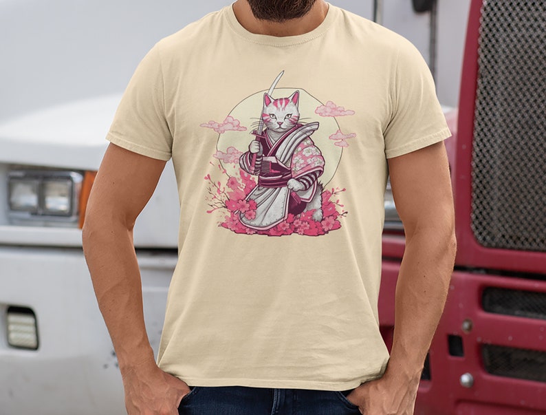 K&ouml;nnte beinhalten: Ein beiges T-Shirt mit einem Aufdruck eines wei&szlig;en Katers, der ein rosa-violettes Samurai-Outfit tr&auml;gt und ein Schwert h&auml;lt. Die Katze steht vor einem rosa Kirschbl&uuml;tenbaum und einem Vollmond.
