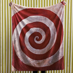 May include: A red and white spiral patterned blanket. The blanket is held up against a yellow and white striped background. The spiral design is centered on the blanket, with a distressed, vintage look.