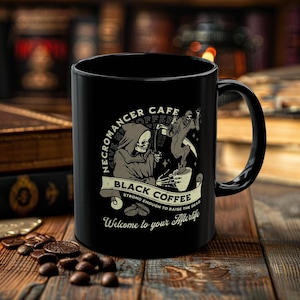 May include: Black coffee mug with a black and white illustration of a skeleton in a hooded robe holding a coffee cup. The text on the mug reads "Necromancer Cafe Give Me My Coffee Black Coffee Strong Enough To Raise The Dead Welcome To Your Afterlife".