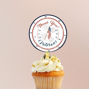 May include: A cupcake topped with white frosting and gold sprinkles with a round, white and blue paper topper that says "Thank You! Veterans" with a red, white, and blue American flag and fireworks.
