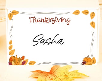 Puede incluir: Una tarjeta de mesa blanca de Acción de Gracias con el nombre "Sasha" en escritura negra. La tarjeta está decorada con calabazas naranjas, hojas de otoño y la palabra "Thanksgiving" en escritura marrón. Una hoja decorativa descansa sobre el plato.