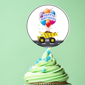 May include: A green cupcake with blue star sprinkles topped with a round cake topper featuring a yellow dump truck carrying colorful balloons. The cake topper says "Happy Birthday".
