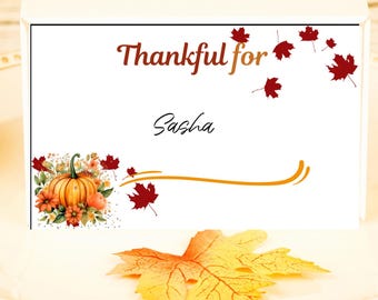Puede incluir: Una tarjeta de mesa blanca con el texto "Thankful for Sasha" en una fuente decorativa, adornada con ilustraciones otoñales de calabazas y hojas de arce. Una hoja amarilla y naranja descansa sobre un plato blanco.
