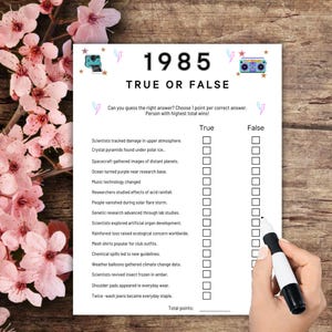 Puede incluir: Una hoja de juego "1985 Verdadero o Falso" en blanco y negro con preguntas de opción múltiple. Una mano con un marcador negro está a punto de seleccionar una respuesta. La hoja está sobre una superficie de madera, con flores rosas en el fondo.