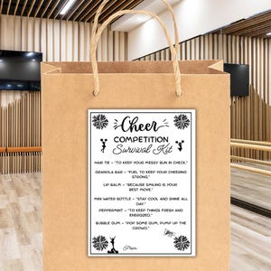 Könnte beinhalten: Eine braune Papiertüte mit einer weißen Karte mit der Aufschrift "Cheer Competition Survival Kit". Das Set enthält ein Haargummi, einen Müsliriegel, Lippenbalsam, eine Mini-Wasserflasche, Pfefferminze und Kaugummi. Die Tüte hat einen gedrehten Papiergriff.