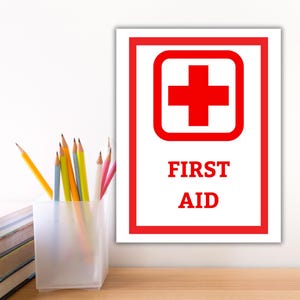 Puede incluir: Un letrero blanco con un borde rojo y un símbolo de cruz roja dentro de un cuadrado rojo. Las palabras "FIRST AID" están impresas en rojo debajo de la cruz. Se ven lápices en un recipiente transparente y una pila de libros.
