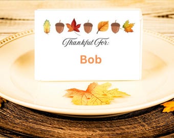 Puede incluir: Una tarjeta blanca con el texto "Thankful For: Bob" e ilustraciones de hojas de otoño y bellotas. La tarjeta está sobre un plato blanco con borde dorado, con hojas de otoño esparcidas.