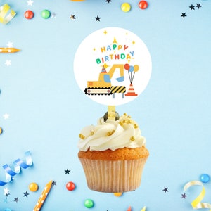 May include: A cupcake with white frosting and gold sprinkles, topped with a round "Happy Birthday" cake topper featuring a yellow excavator, balloons, and a construction cone. The background is light blue with party decorations.