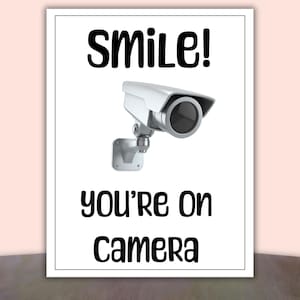 May include: A white framed print with the words "Smile! You're On Camera" in black text. A security camera is depicted in the center of the print. The camera is silver and black.