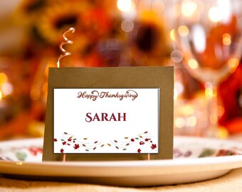 Puede incluir: Una tarjeta de mesa de Acción de Gracias con el nombre "SARAH" en escritura burdeos. La tarjeta está colocada sobre un plato blanco con un diseño floral. Las palabras "Happy Thanksgiving" están encima del nombre, y la tarjeta está decorada con hojas de otoño.
