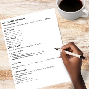 May include: A white document titled "CATTLE LEASE AGREEMENT" with fill-in-the-blank sections for Lessor and Lessee details. A hand holding a pen is poised to write on the document. A white coffee cup sits nearby on a wooden surface.