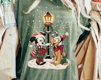 May include: Green long-sleeved t-shirt with a Christmas-themed design featuring Mickey Mouse and Pluto standing in front of a street lamp with a wreath. The text "Merry Christmas" is written above the characters.