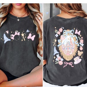May include: Dark grey t-shirt featuring a whimsical design with a magic wand, Minnie Mouse ears, butterflies, and a wizard hat on the front. The back showcases a castle within a decorative frame, surrounded by flowers and bows.