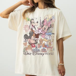 May include: An off-white short-sleeve t-shirt featuring a graphic print of Mickey Mouse, Minnie Mouse, Goofy, and Donald Duck. The design includes various Walt Disney World park landmarks, fireworks, and the text Walt Disney World in a script font.