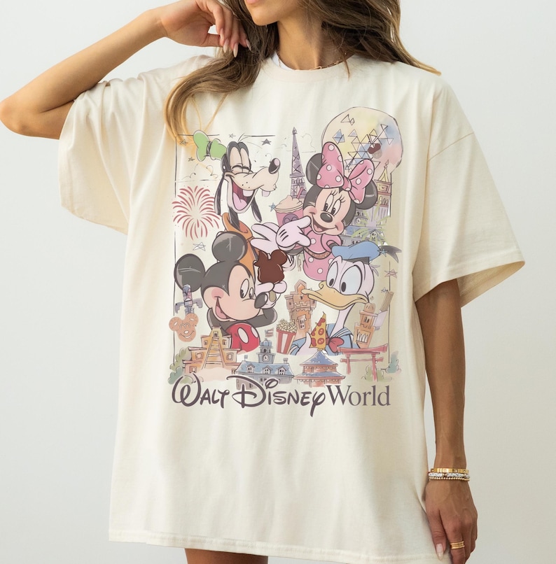 May include: An off-white short-sleeve t-shirt featuring a graphic print of Mickey Mouse, Minnie Mouse, Goofy, and Donald Duck. The design includes various Walt Disney World park landmarks, fireworks, and the text Walt Disney World in a script font.