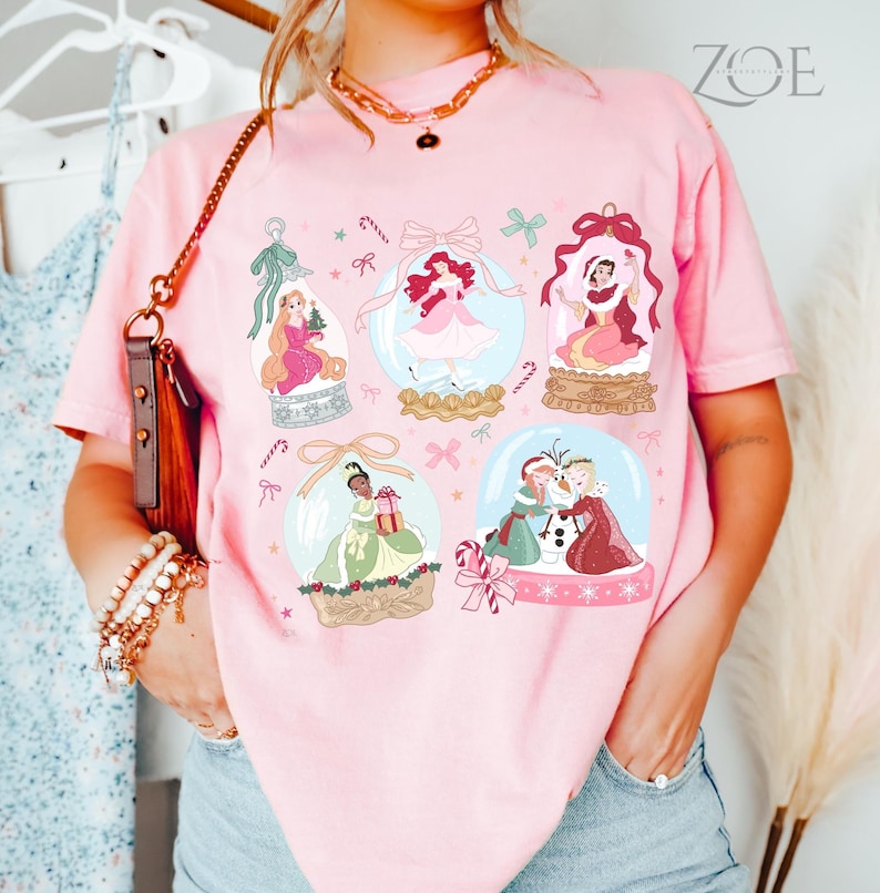 May include: Pink t-shirt featuring illustrations of princesses inside snow globes. The design includes candy canes, bows, and snowflakes. The text "ZOE" is printed in the upper right corner.