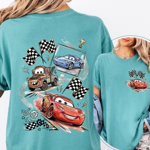Puede incluir: Camiseta de color turquesa con un dise&ntilde;o de coches de carreras de dibujos animados, incluyendo un coche rojo, una gr&uacute;a marr&oacute;n y un coche azul, con banderas a cuadros y un trofeo. Una versi&oacute;n m&aacute;s peque&ntilde;a del dise&ntilde;o est&aacute; en el bolsillo.