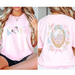 May include: A light pink t-shirt featuring a cartoon design of a mouse with a bow, a wizard hat, a wand, and a bow. The back of the shirt has a castle design inside a gold frame with flowers and bows.