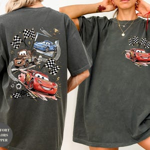 Puede incluir: Camiseta gris oscuro con un gr&aacute;fico de Rayo McQueen, Mate y otros personajes de Cars. La camiseta tiene un aspecto desgastado e incluye banderas a cuadros y un trofeo. El texto "COMFORT COLORS PEPPER" est&aacute; impreso en la parte inferior izquierda.