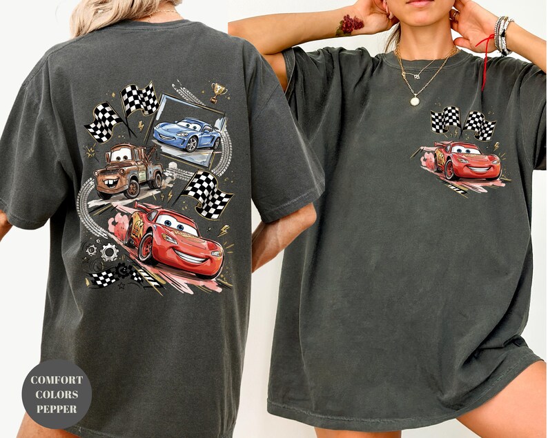 Puede incluir: Camiseta gris oscuro con un gr&aacute;fico de Rayo McQueen, Mate y otros personajes de Cars. La camiseta tiene un aspecto desgastado e incluye banderas a cuadros y un trofeo. El texto "COMFORT COLORS PEPPER" est&aacute; impreso en la parte inferior izquierda.