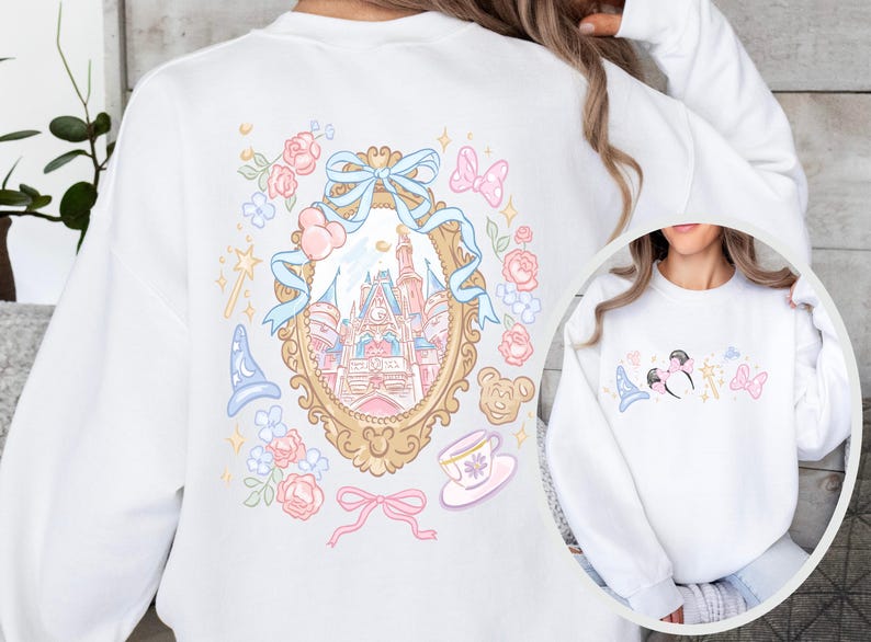 May include: White sweatshirt featuring a whimsical illustration of a castle within a gold-framed mirror, surrounded by pastel-colored flowers, bows, and a magic wand. A smaller, matching design is on the front. The design is reminiscent of a fairytale theme.