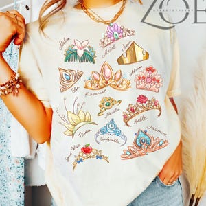 K&ouml;nnte beinhalten: Cremefarbenes T-Shirt mit farbenfrohen Illustrationen verschiedener Prinzessinnen-Diademe, die jeweils mit einem Namen versehen sind. Das Design umfasst Diademe f&uuml;r Arielle, Aurora, Belle, Cinderella, Elsa, Jasmin, Mulan, Moana, Rapunzel, Tiana und Schneewittchen.