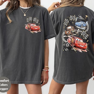 Puede incluir: Camiseta gris oscuro con un coche de carreras de dibujos animados rojo y banderas a cuadros en la parte delantera. La parte trasera de la camiseta muestra tres coches de carreras de dibujos animados, banderas a cuadros y un trofeo. La camiseta est&aacute; hecha de un material suave y c&oacute;modo.