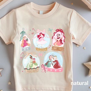 May include: A natural-colored t-shirt featuring six snow globes with princess illustrations. Each globe has a different princess, with candy cane and bow accents. The word "natural" is printed on the bottom right.