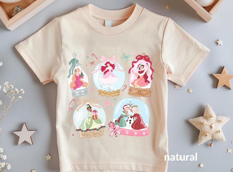 May include: A natural-colored t-shirt featuring six snow globes with princess illustrations. Each globe has a different princess, with candy cane and bow accents. The word "natural" is printed on the bottom right.