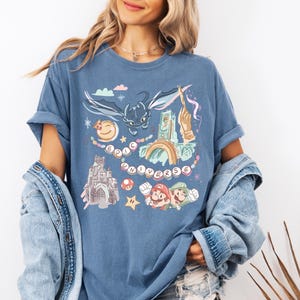 K&ouml;nnte beinhalten: Ein blaues T-Shirt mit einem grafischen Druck eines Drachen, einer Burg und Figuren aus beliebten Videospielen. Der Text "EPIC UNIVERSE" ist auf dem T-Shirt gedruckt.