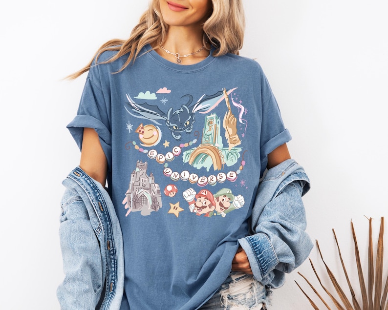 K&ouml;nnte beinhalten: Ein blaues T-Shirt mit einem grafischen Druck eines Drachen, einer Burg und Figuren aus beliebten Videospielen. Der Text "EPIC UNIVERSE" ist auf dem T-Shirt gedruckt.