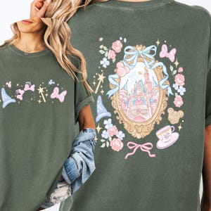 May include: Olive green t-shirt featuring a whimsical design with a castle, bows, and floral elements. The front displays a wizard hat, Minnie Mouse ears, and butterflies. The back showcases a castle within a decorative frame.
