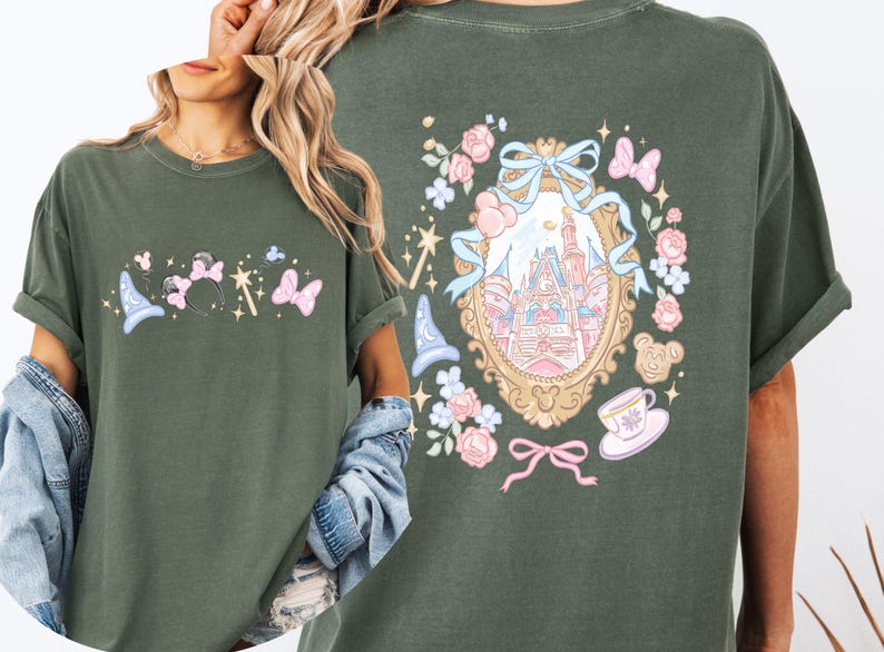May include: Olive green t-shirt featuring a whimsical design with a castle, bows, and floral elements. The front displays a wizard hat, Minnie Mouse ears, and butterflies. The back showcases a castle within a decorative frame.