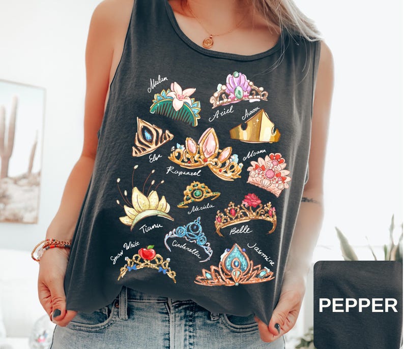 May include: Dark gray tank top featuring colorful illustrations of various princess tiaras. Each tiara is labeled with a princess name, including Ariel, Belle, and Jasmine. The tank top is made of a soft material and is perfect for casual wear.