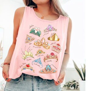 May include: Pink tank top featuring illustrations of various princess crowns with names like Ariel, Belle, and Jasmine. The crowns are in different colors and styles, with details like gems and flowers. The tank top is made of a soft material.
