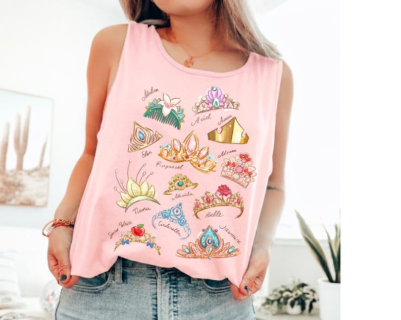 May include: Pink tank top featuring illustrations of various princess crowns with names like Ariel, Belle, and Jasmine. The crowns are in different colors and styles, with details like gems and flowers. The tank top is made of a soft material.