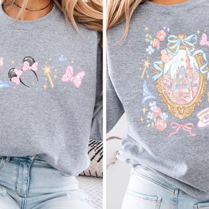 May include: A gray sweatshirt featuring a whimsical design. The front showcases a blue wizard hat, Minnie Mouse ears with a pink bow, a golden wand, and a pink butterfly. The back displays a castle within a gold frame, surrounded by flowers and bows.