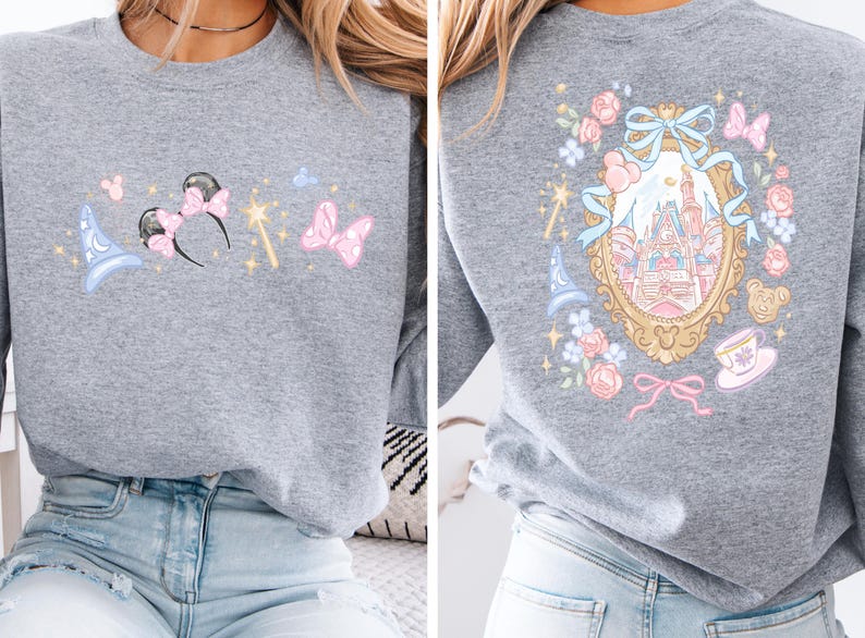 May include: A gray sweatshirt featuring a whimsical design. The front showcases a blue wizard hat, Minnie Mouse ears with a pink bow, a golden wand, and a pink butterfly. The back displays a castle within a gold frame, surrounded by flowers and bows.