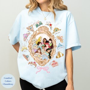 May include: Light blue t-shirt featuring a framed illustration of Disney princesses. Surrounding the frame are various princess tiaras and crowns. The text "Zoe Z" is visible. The shirt also has the text "Comfort Colors Chambray".