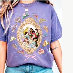 May include: A purple t-shirt featuring a framed illustration of Disney princesses. The shirt has a vintage look, with various princess tiaras and the word "Zoe" printed around the frame. The shirt is worn with blue jeans.