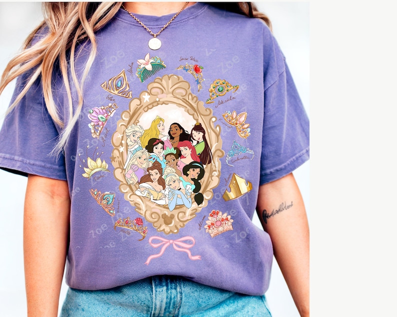 May include: A purple t-shirt featuring a framed illustration of Disney princesses. The shirt has a vintage look, with various princess tiaras and the word "Zoe" printed around the frame. The shirt is worn with blue jeans.