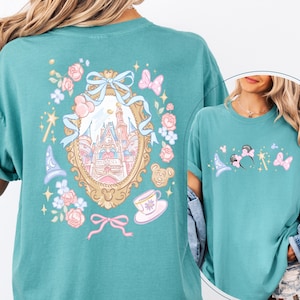 May include: Teal t-shirt featuring a whimsical design with a castle, flowers, and butterflies. The front of the shirt has a smaller design with a magic wand, mouse ears, and a wizard hat. The shirt has a relaxed fit.