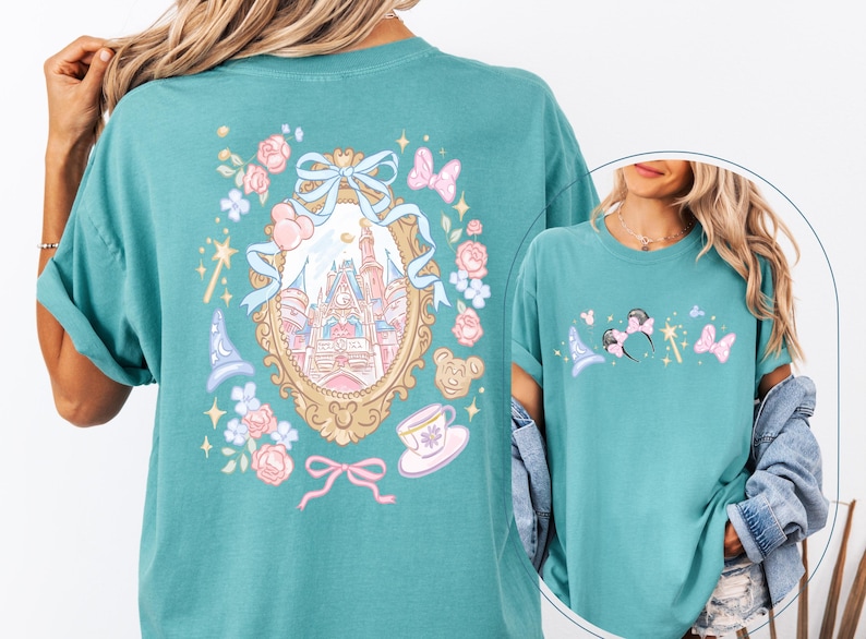 May include: Teal t-shirt featuring a whimsical design with a castle, flowers, and butterflies. The front of the shirt has a smaller design with a magic wand, mouse ears, and a wizard hat. The shirt has a relaxed fit.