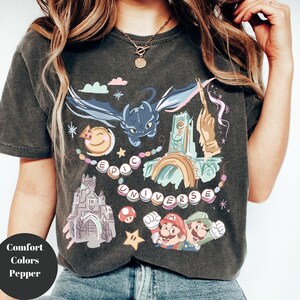 K&ouml;nnte beinhalten: Ein dunkelgraues T-Shirt mit einem bunten Grafikdruck, der einen Drachen, ein Schloss, einen Regenbogen, einen Pilz und die Figuren Mario und Luigi zeigt. Der Text "EPIC UNIVERSE" ist in einem Kreis um die Grafik gedruckt.