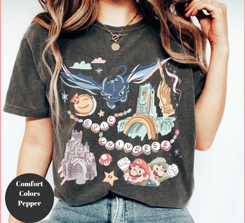 K&ouml;nnte beinhalten: Ein dunkelgraues T-Shirt mit einem bunten Grafikdruck, der einen Drachen, ein Schloss, einen Regenbogen, einen Pilz und die Figuren Mario und Luigi zeigt. Der Text "EPIC UNIVERSE" ist in einem Kreis um die Grafik gedruckt.